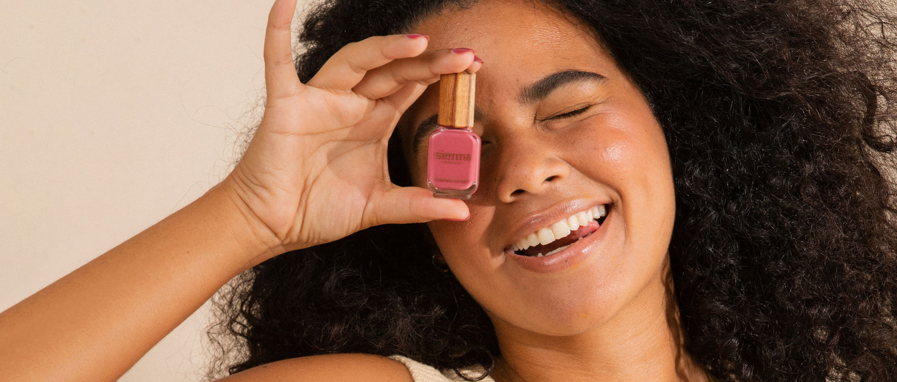 dark haired woman wearing pink nail polish and covering one eye with nail polish bottle