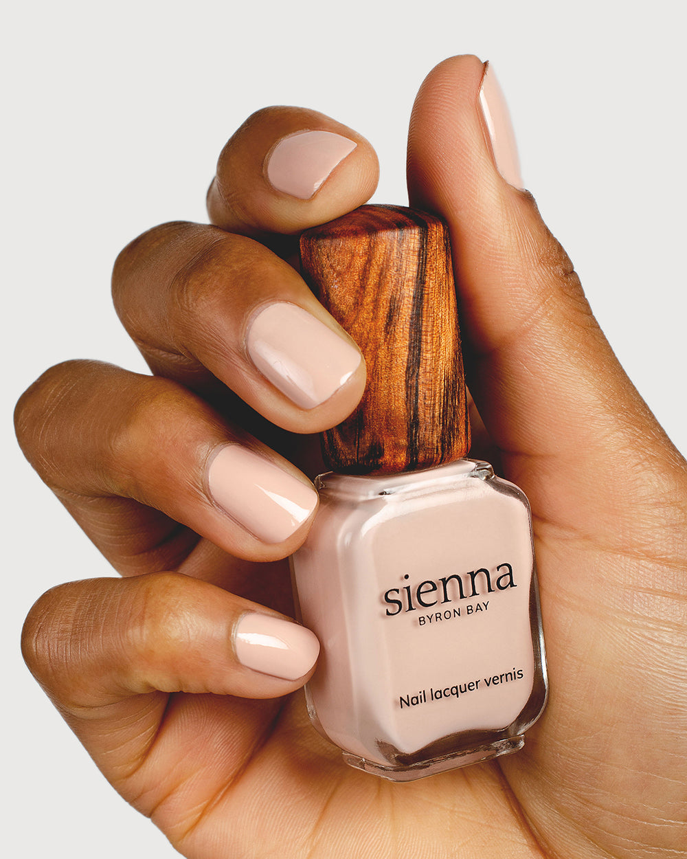 Soft neutral-pink nail polish hand swatch on medium skin tone holding sienna bottle close-up