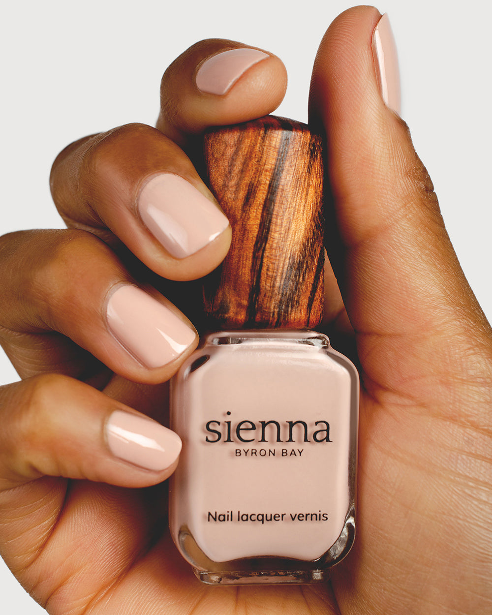 Soft neutral-pink nail polish hand swatch on medium skin tone holding sienna bottle close-up